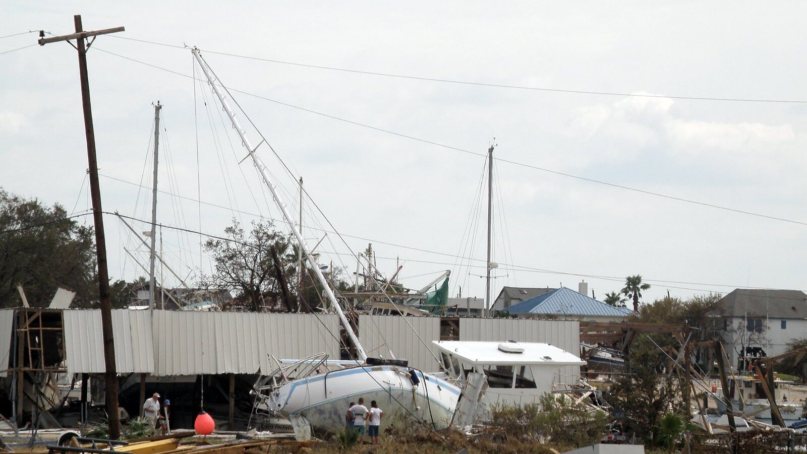 Image of a boat stranded on land, leaning against a wind-destroyed structure and a power line, amid other debris, including destroyed buildings and cars. Three people stand next to the boat observing the damage.