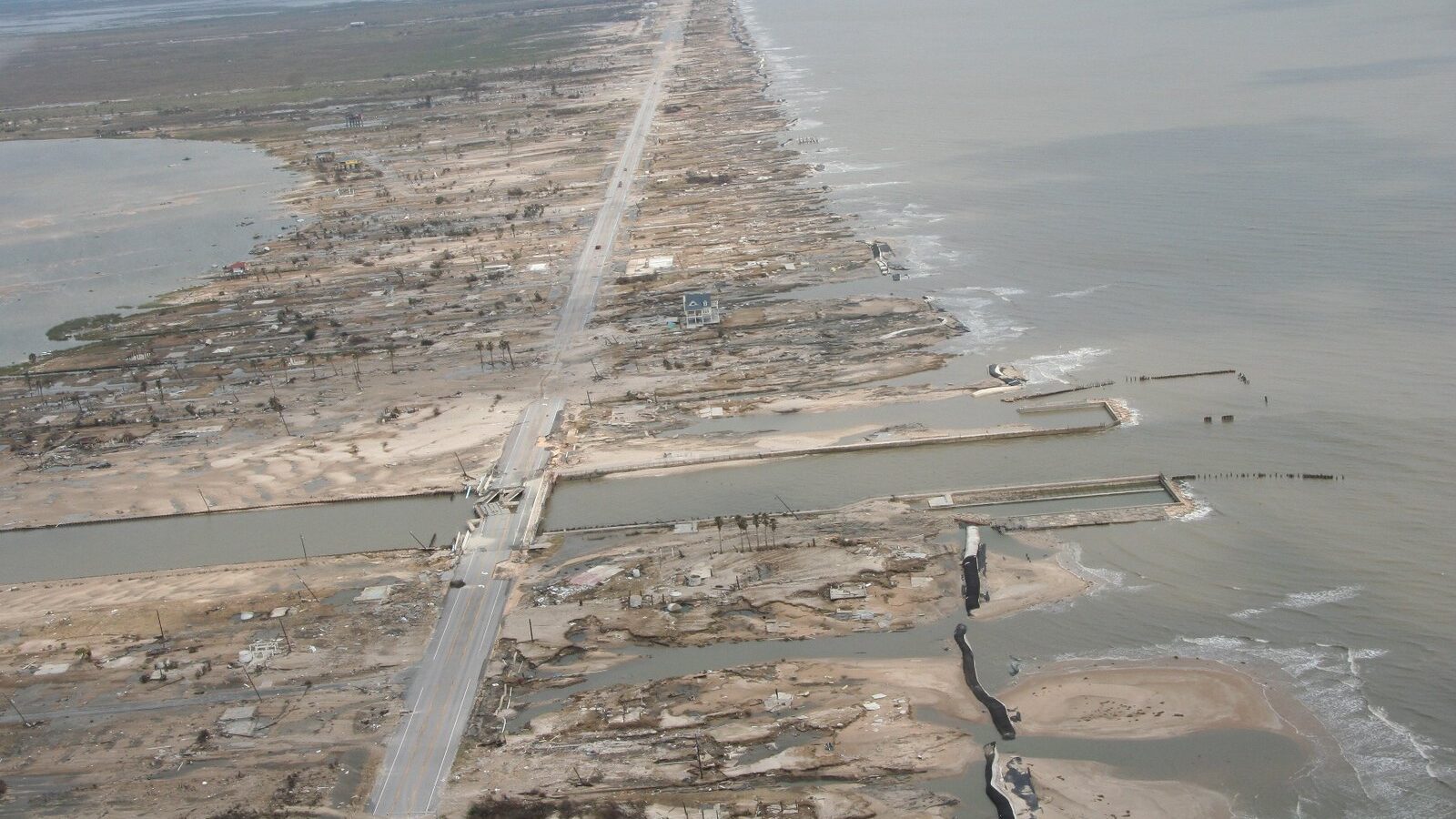 Aerial photo of a peninsula/barrier island that has been breached by a storm surge. Sand, buildings, and other structures have been washed away or damaged, including visible broken lines of sandbags, a damaged bridge, damaged trees, and obliterated buildings; only one house appears to still be standing.
