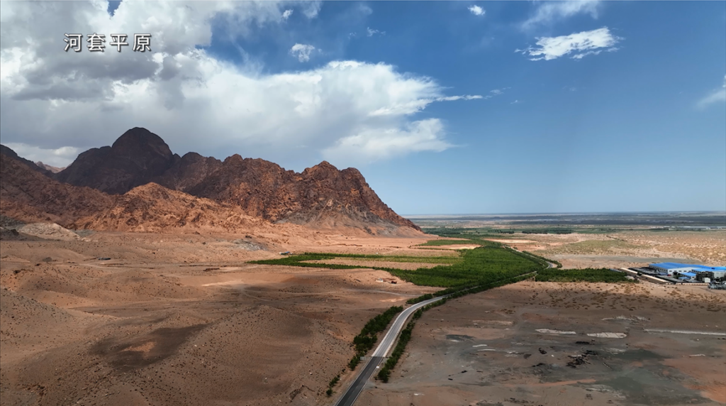 Image showing treeless mountains and sparsely vegetated foothills next to flat land. In the center of the photo is a dark green area of vegetation which contrasts sharply with the otherwise brown/tan landscape. A road runs to one edge of the green area. At the very right of the image, in the middle distance, is a large, wide building with a bright blue roof.