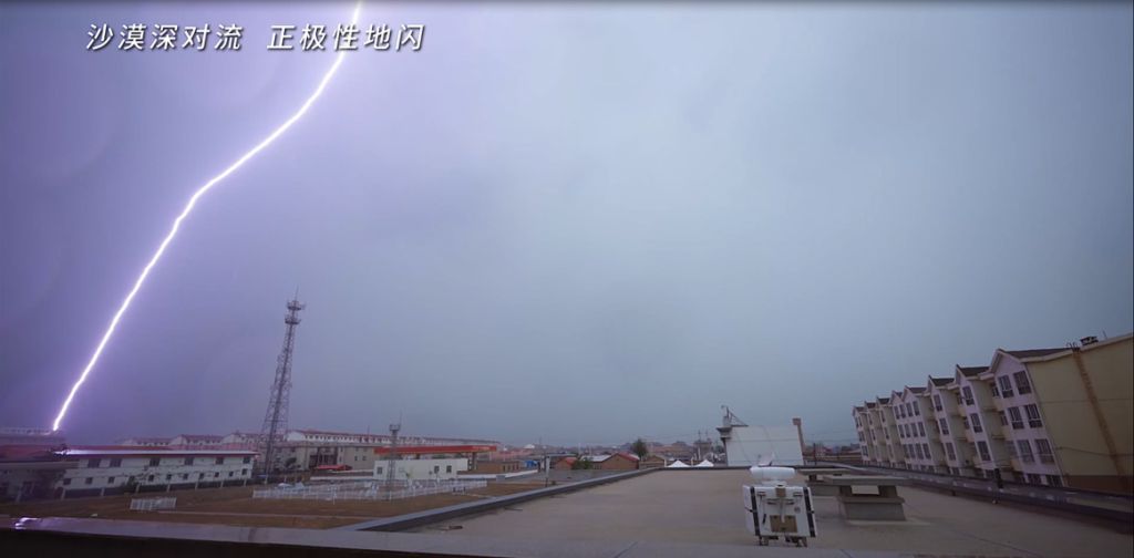 Video still shows a bright bolt of lightning in the far left of the image. In the bottom right, a laser wind LIDAR device sits on a rooftop, pointing in the direction of the storm.