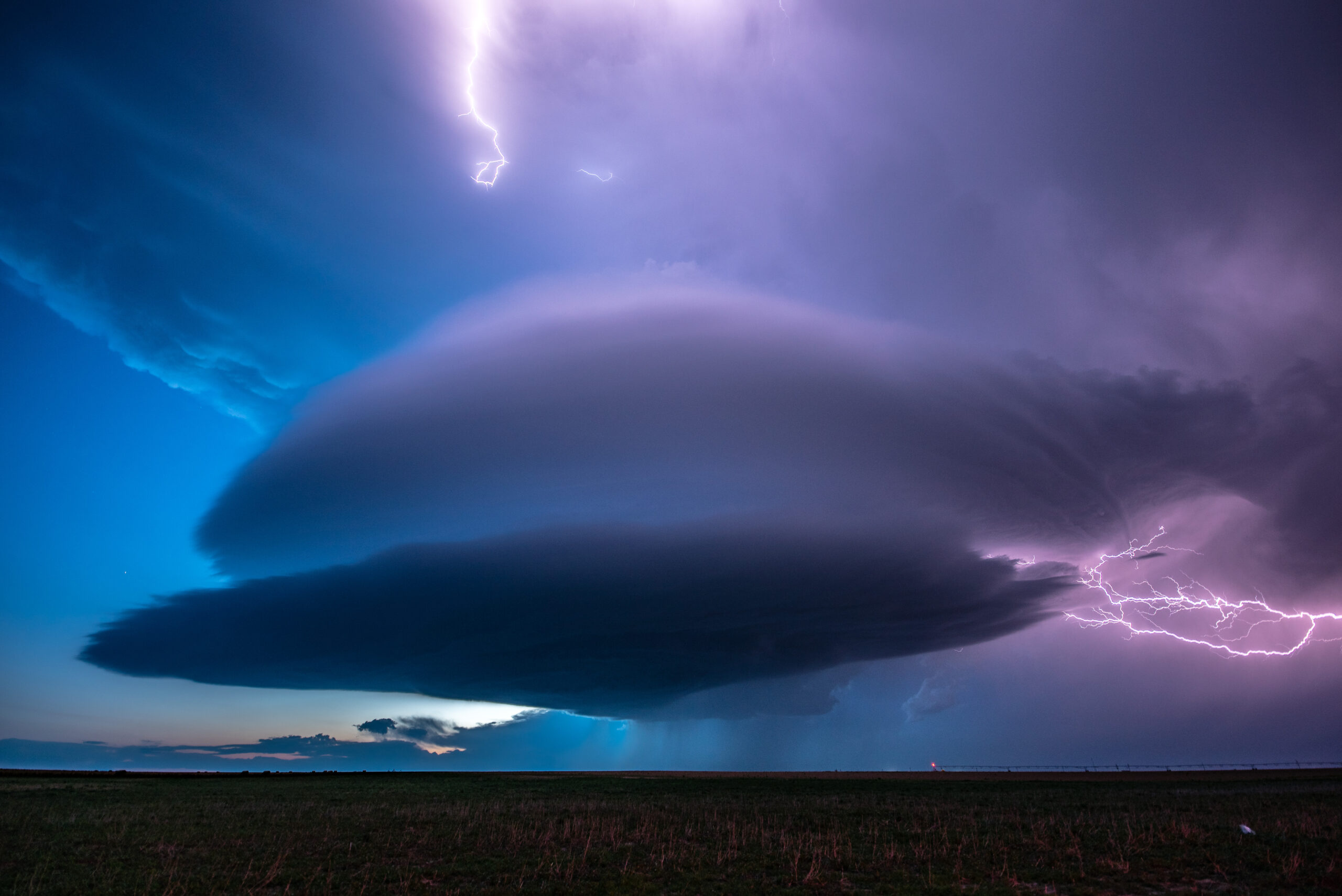 Photo "Walsh Mothership" by Michael Seger shows a rounded storm from a distance at twilight, with a lightning flash in the sky in the foreground. The photo was voted Member's Choice in the 2023 AMS Weather Band Photo Contest.